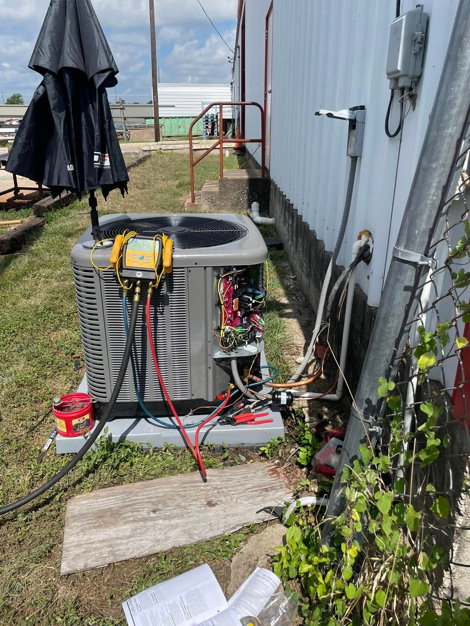 AC unit being serviced outdoors, hoses and gauges attached, electrical box nearby, paperwork.