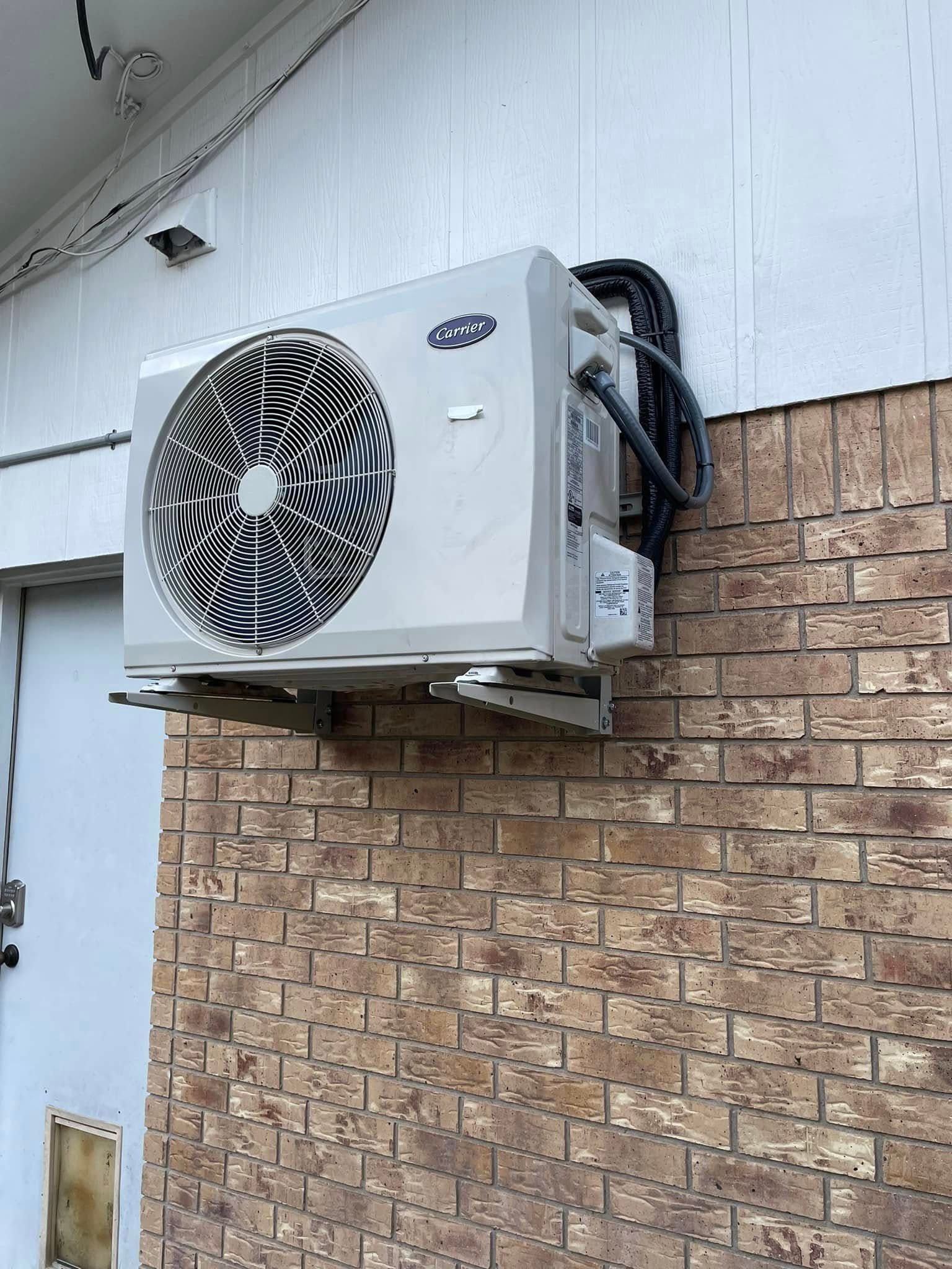 Wall-mounted air conditioner unit on a brick exterior wall. Black power cord and tubing run along the wall.