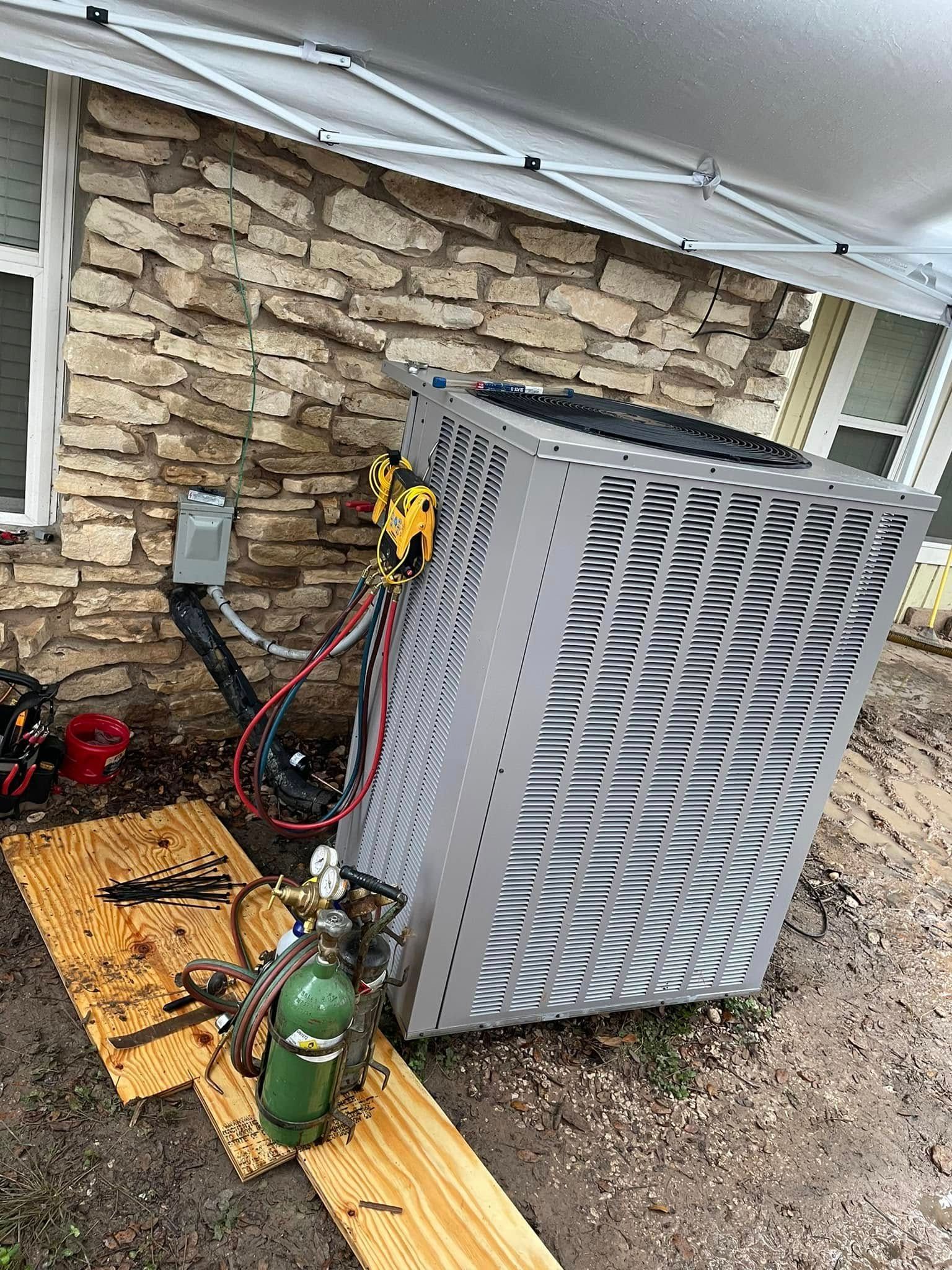 HVAC unit being worked on outdoors; near a brick wall, with tools and a gas tank.