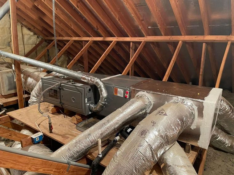 HVAC unit in an attic with ductwork, set among wooden beams and insulation.