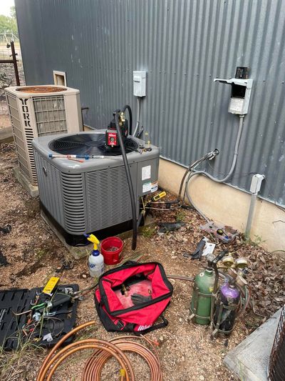 HVAC unit being serviced outdoors, with tools, hoses, and gas cylinders on the ground.