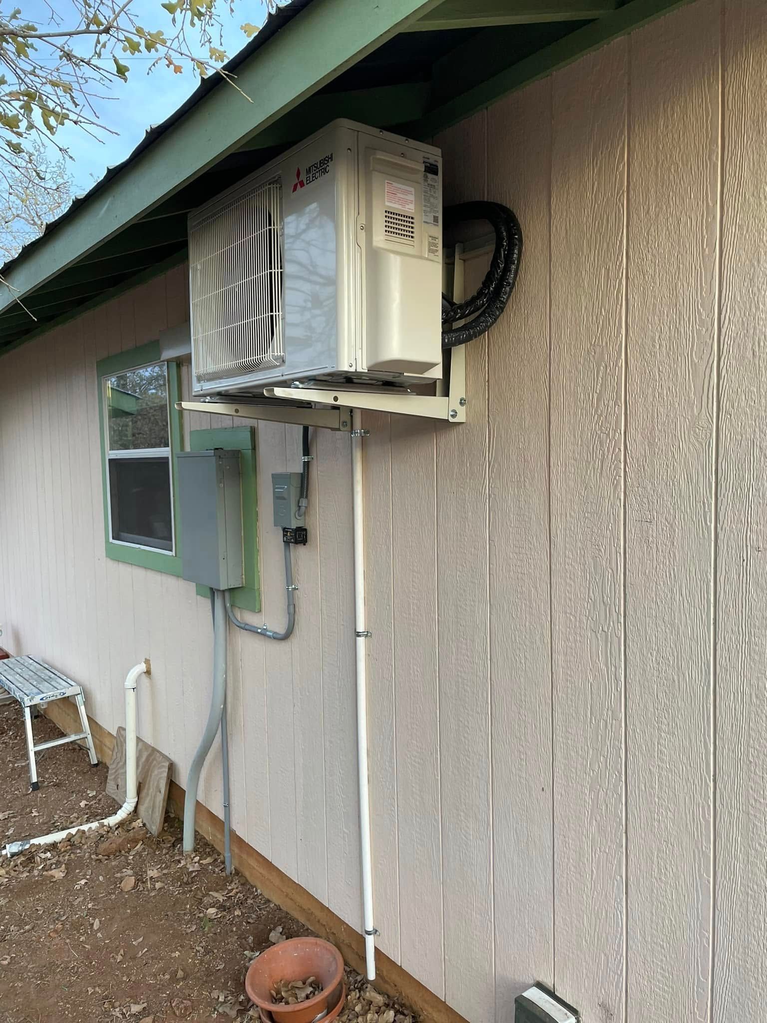 Exterior view of an air conditioning unit mounted on a light-colored building's side wall. Black pipes connect to the unit.