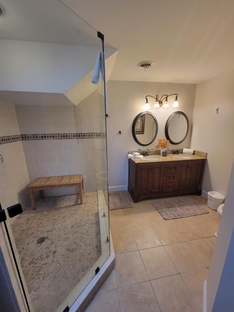Bathroom with glass shower, double vanity with mirrors, and neutral tile flooring.