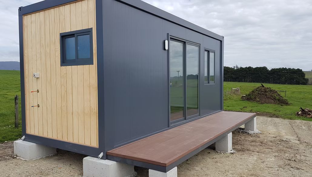 A small, dark gray prefabricated cabin with a light wood-paneled side and a front deck, situated in a grassy field.