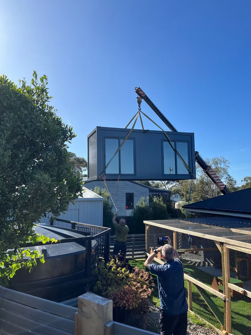 A crane lifts a dark grey modular building unit above a backyard, as two people watch the process from below.