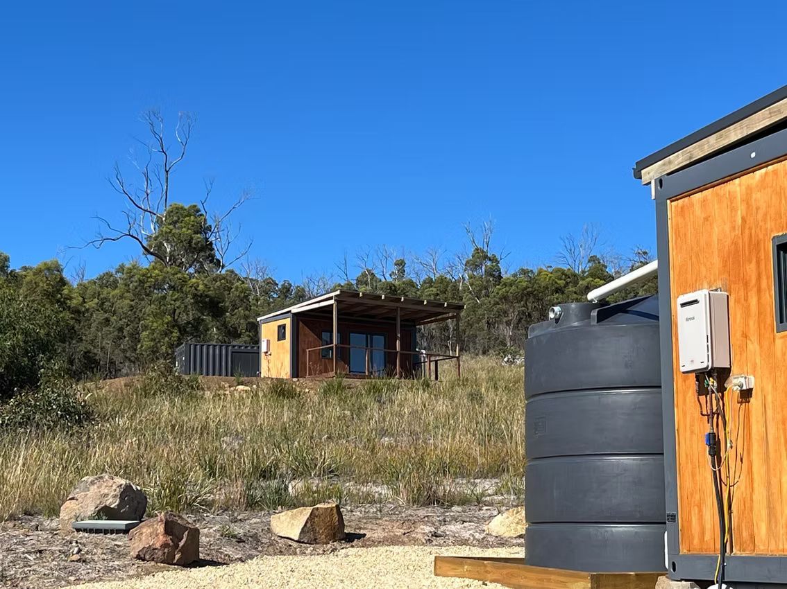 Two small wooden cabins sit on a grassy, sunlit hillside under a clear blue sky, next to a large grey water tank.