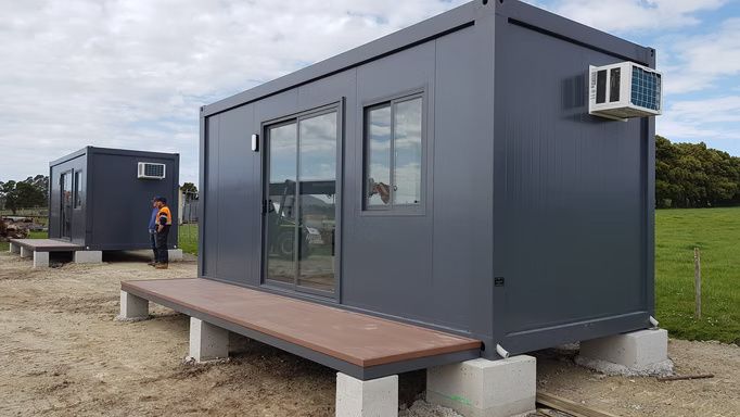 Two dark gray modular shipping container units with sliding doors and air conditioning units, elevated on concrete blocks.