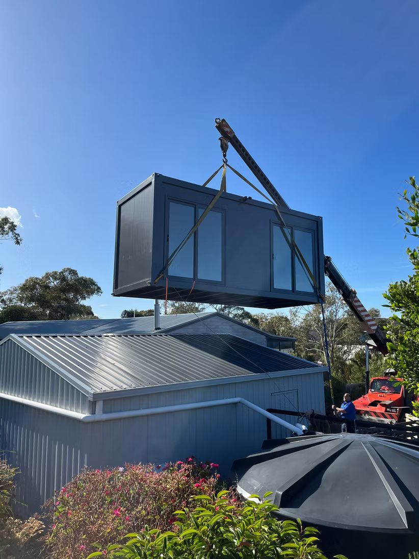 A crane lifts a dark gray modular shipping container over a shed on a sunny day.