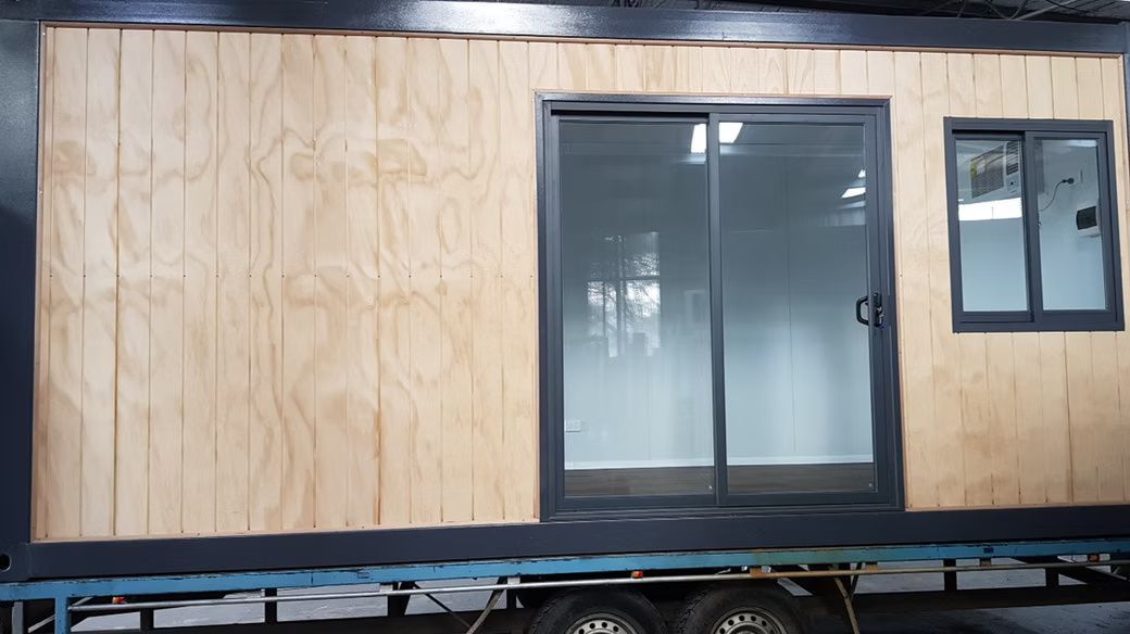 A wooden-paneled trailer structure on wheels featuring a black-framed sliding glass door and a rectangular window.