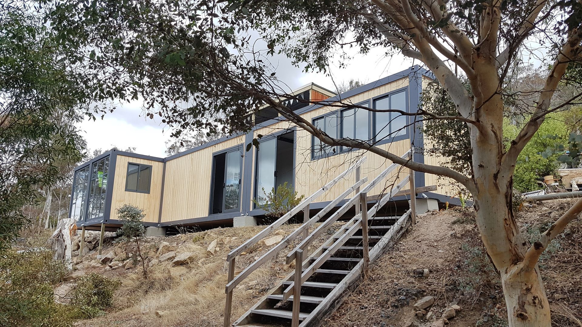 A modular house made of shipping containers sits on a hillside with wooden steps leading up to the entrance.