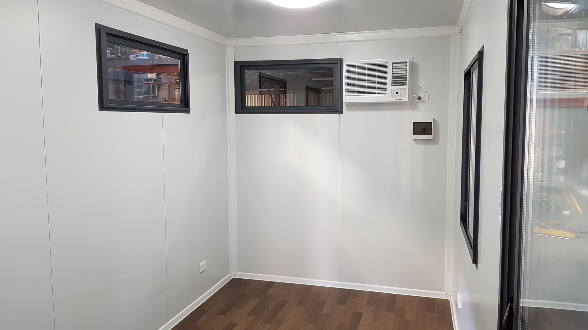 An empty office space with white paneled walls, wood-look flooring, two windows, and a wall-mounted air conditioner.