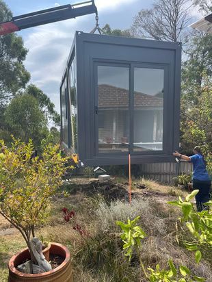 A dark gray modular unit is suspended in the air by a crane, being lowered into a residential yard.