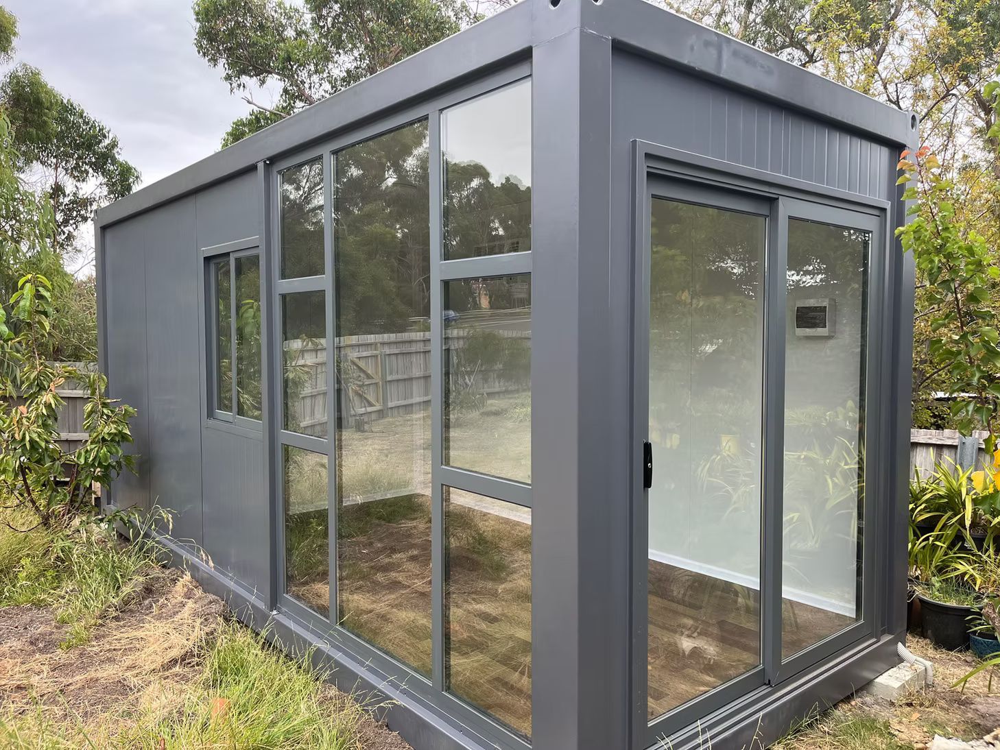 A dark grey, modern prefabricated outdoor pod with large glass walls and sliding doors, set in a grassy backyard.