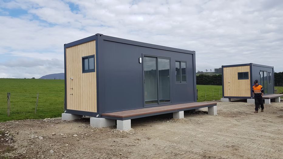 Two modular shipping container homes with wood accents, gray siding, and sliding doors, set on concrete footings in a field.