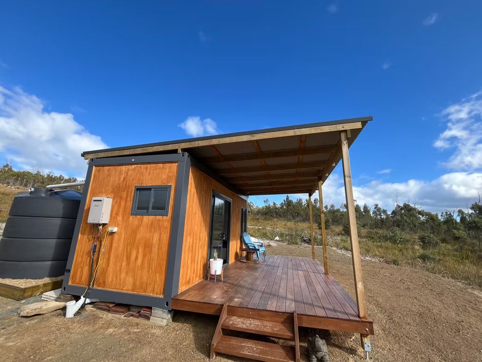 A small cabin made from a shipping container with a wooden deck and porch roof, set in a rural, open landscape.