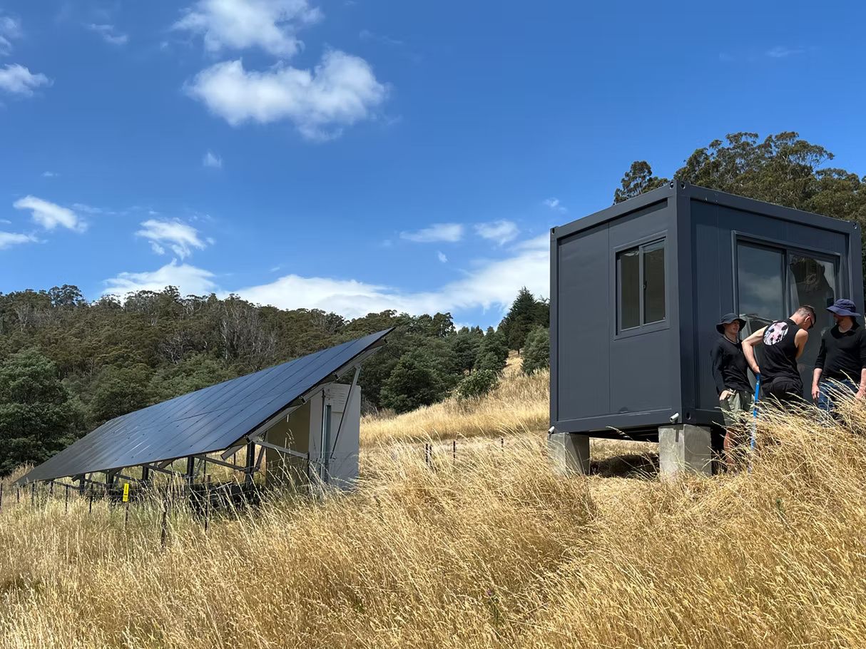 A dark grey, modern modular cabin sits on a grassy hill next to an angled solar panel array under a bright blue sky.