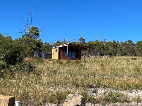 A small, modern wooden cabin with a covered deck sits in a sunny, grassy field surrounded by trees under a clear blue sky.