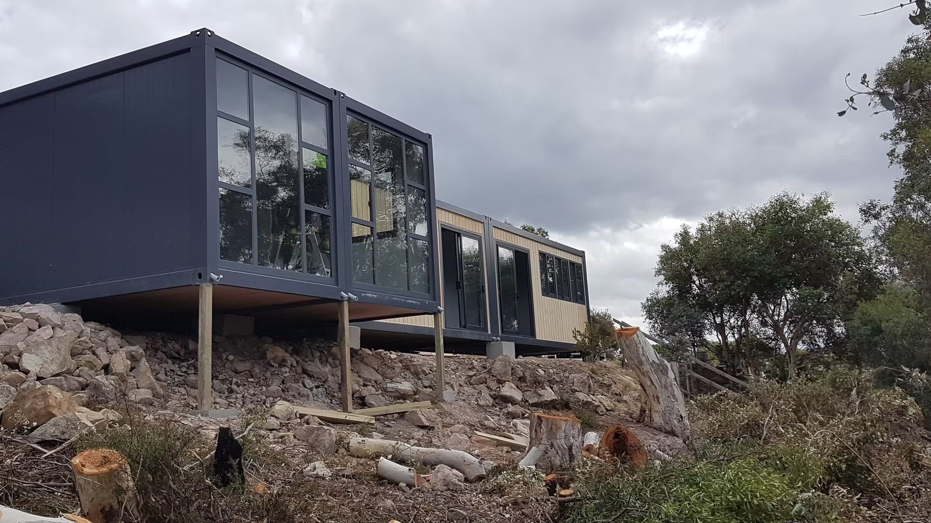 A modern, modular home with large windows, set on a raised platform on a rocky, hillside landscape under a cloudy sky.