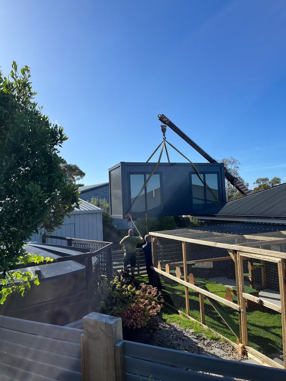 A crane lowers a dark gray modular building unit over a backyard with a chicken coop and foliage on a sunny day.