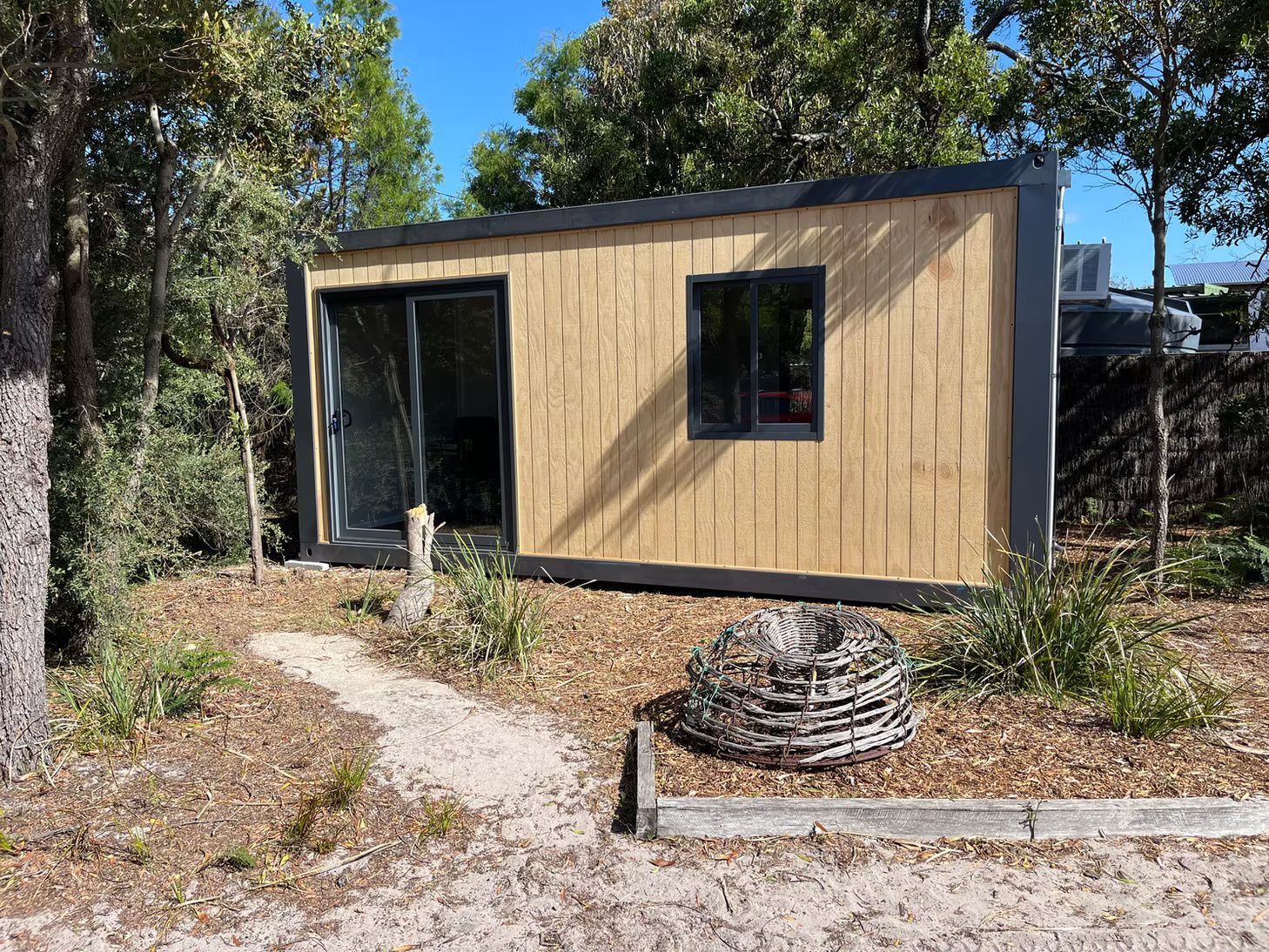 A modern, rectangular cabin with light wood vertical paneling and dark trim, situated in a wooded area with sandy ground.