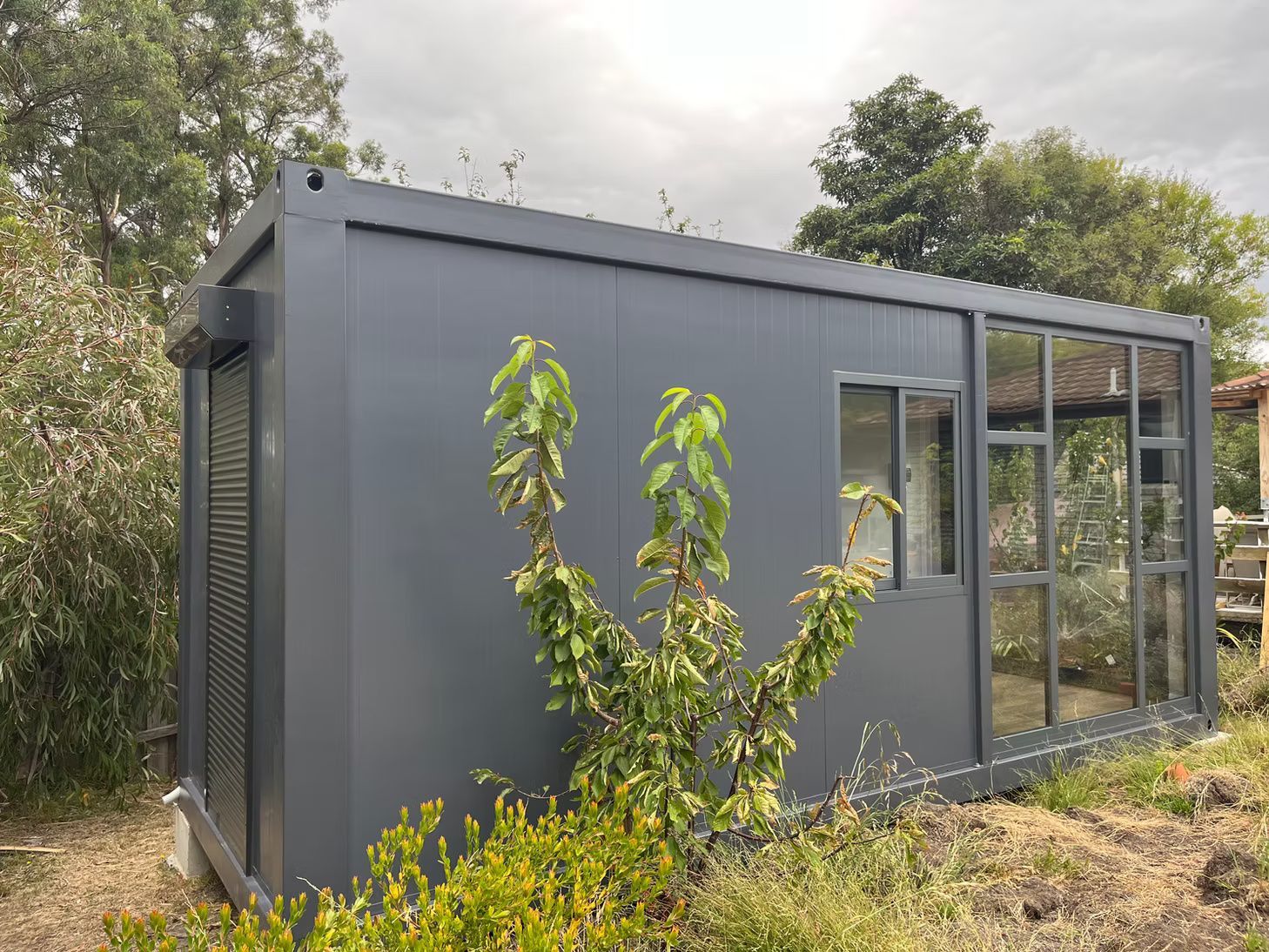 A dark grey portable container office with a window and glass door, situated in an outdoor area with trees and shrubs.