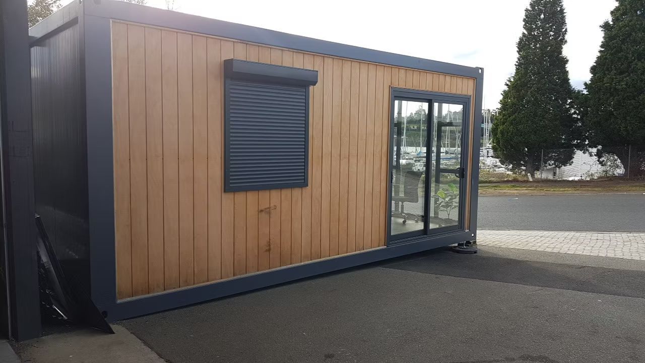 A modern rectangular modular cabin with wood-slat siding, a dark metal frame, a security shuttered window, and glass door.