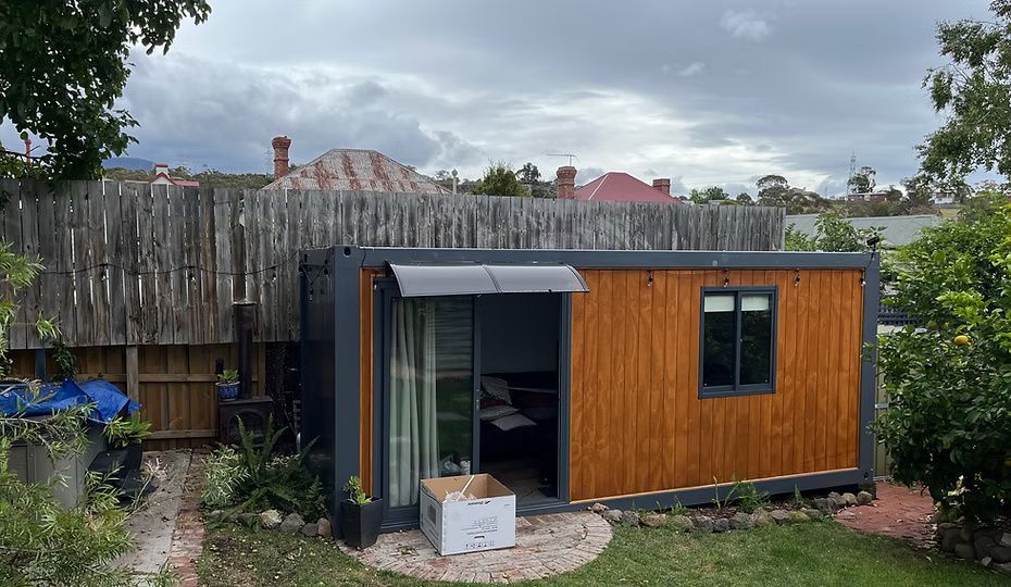 A modern modular studio unit with wood-paneled walls and a sliding glass door, situated in a residential backyard.