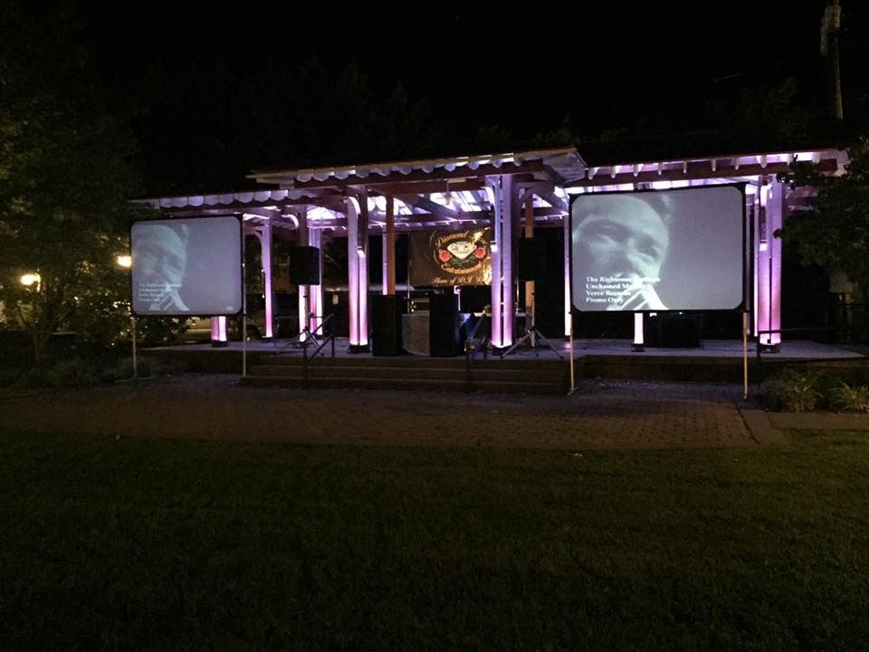 Outdoor nighttime event with a lit stage and two large projection screens in a park