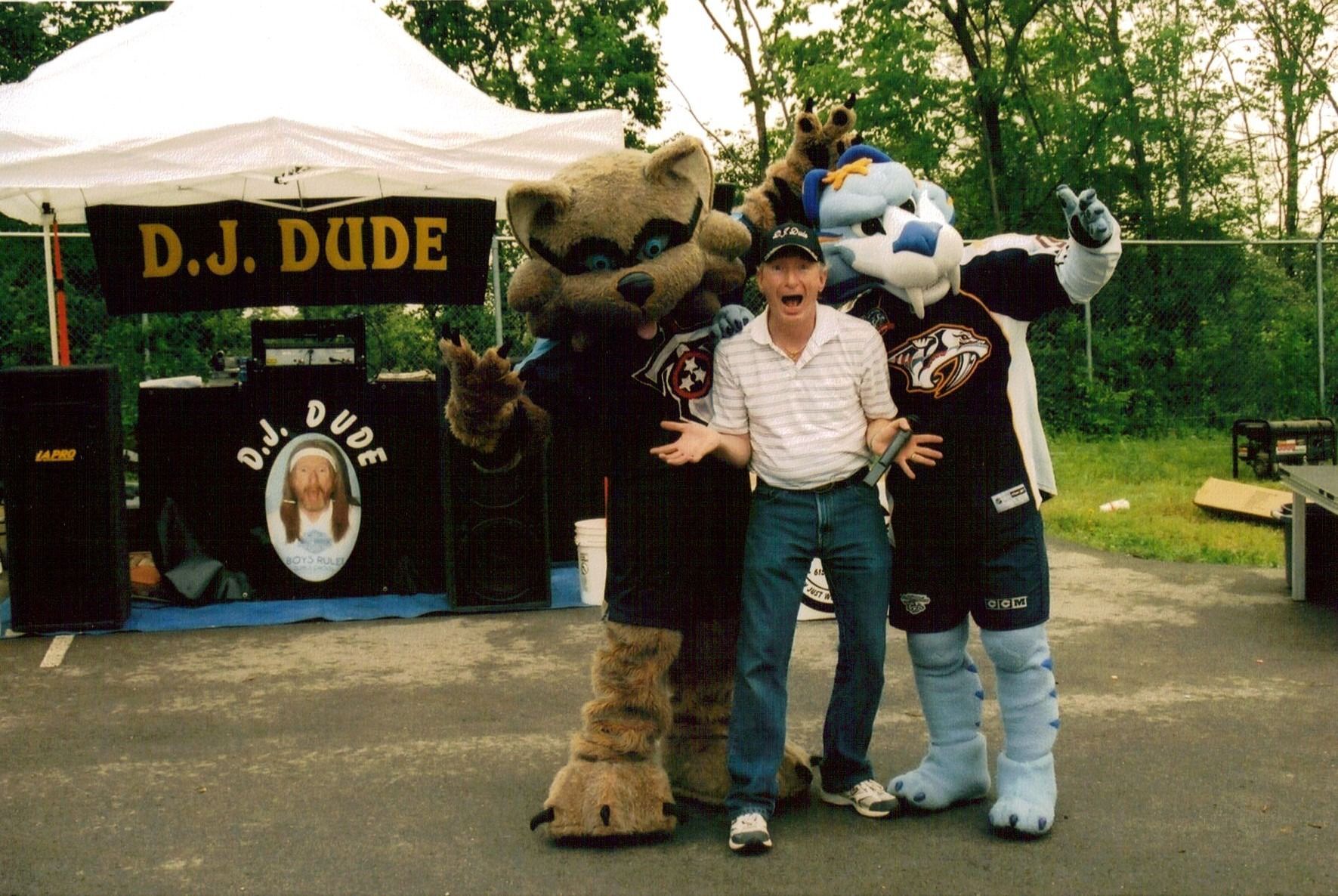 Person posing with two costumed mascots beside a D.J. Duke booth outdoors