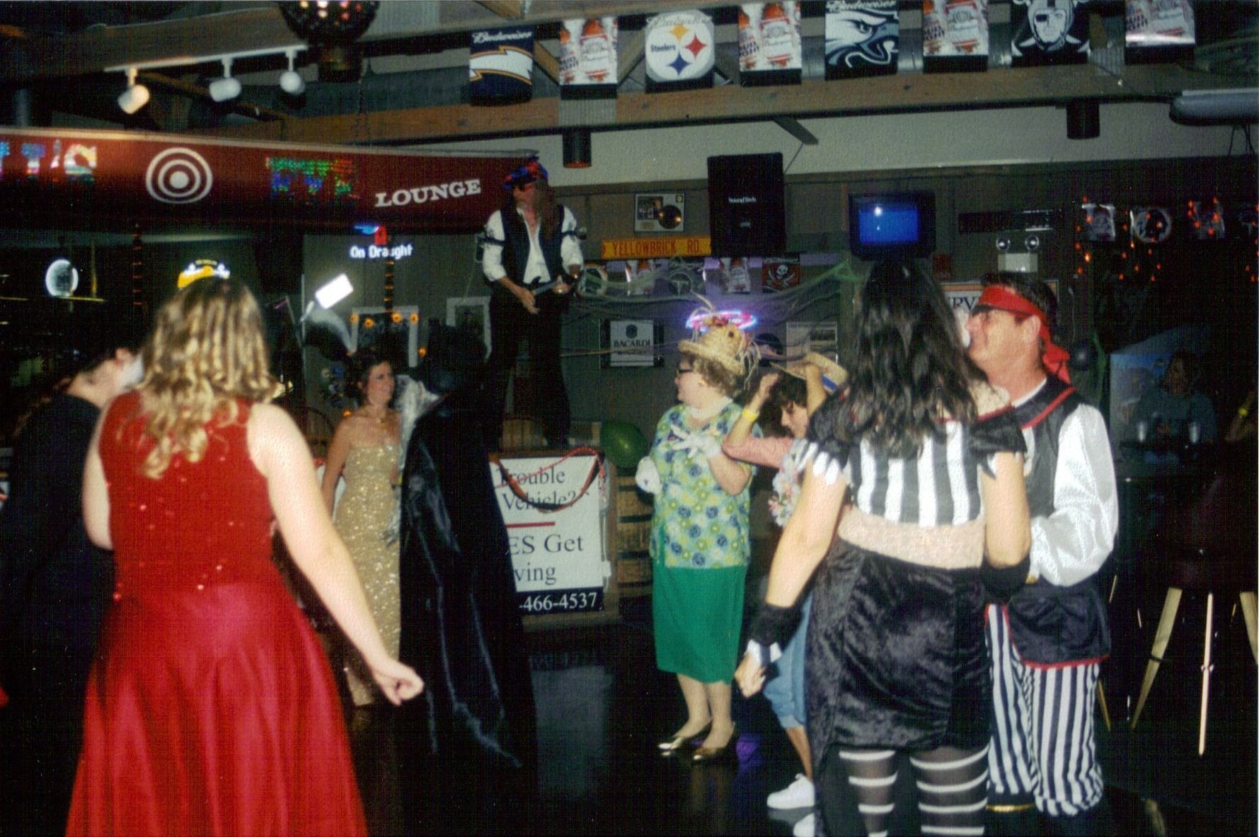People dancing at a crowded bar with colorful costumes and a live performer on stage.