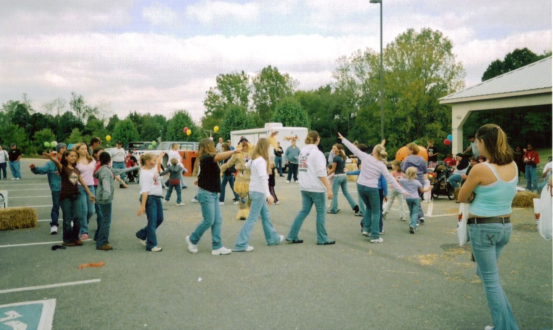 People dancing in a line outdoors near a parking lot and trees, with onlookers nearby