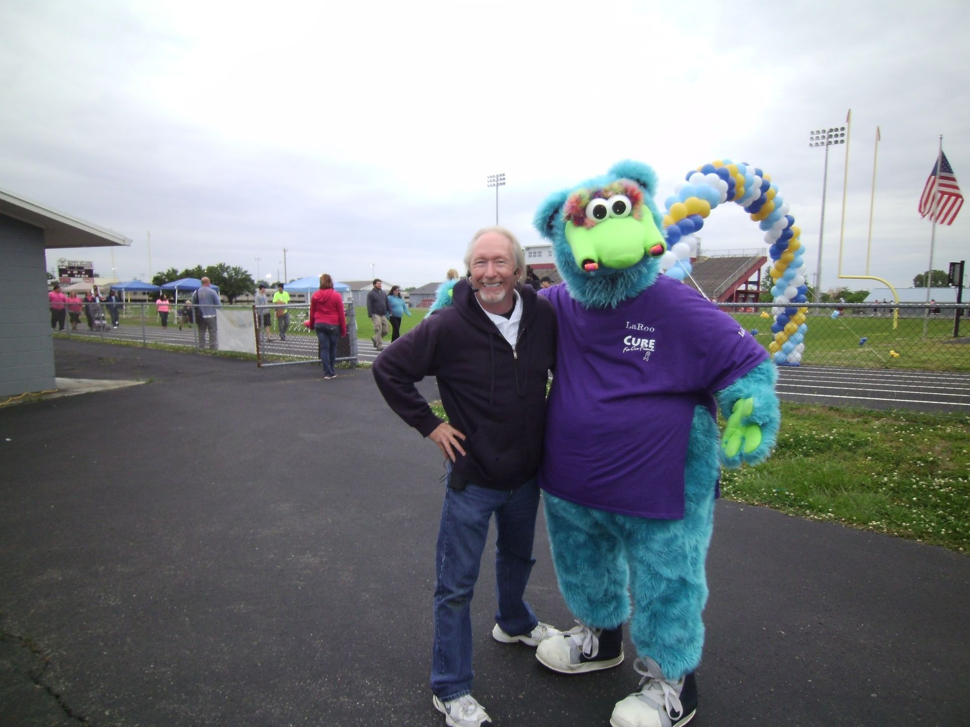 Man and blue furry mascot in purple shirt posing outdoors on a cloudy day