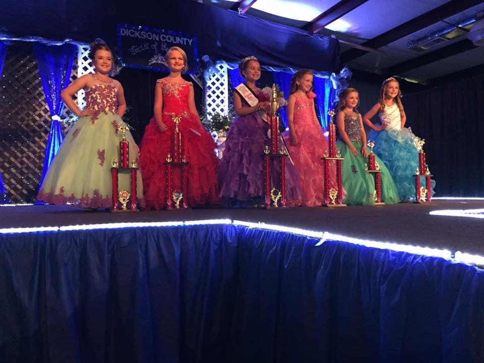 Six women in colorful gowns posing on a lit stage at a pageant event