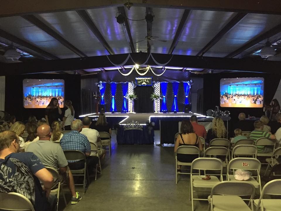 Audience seated in a hall facing a lit stage with screens and blue lights