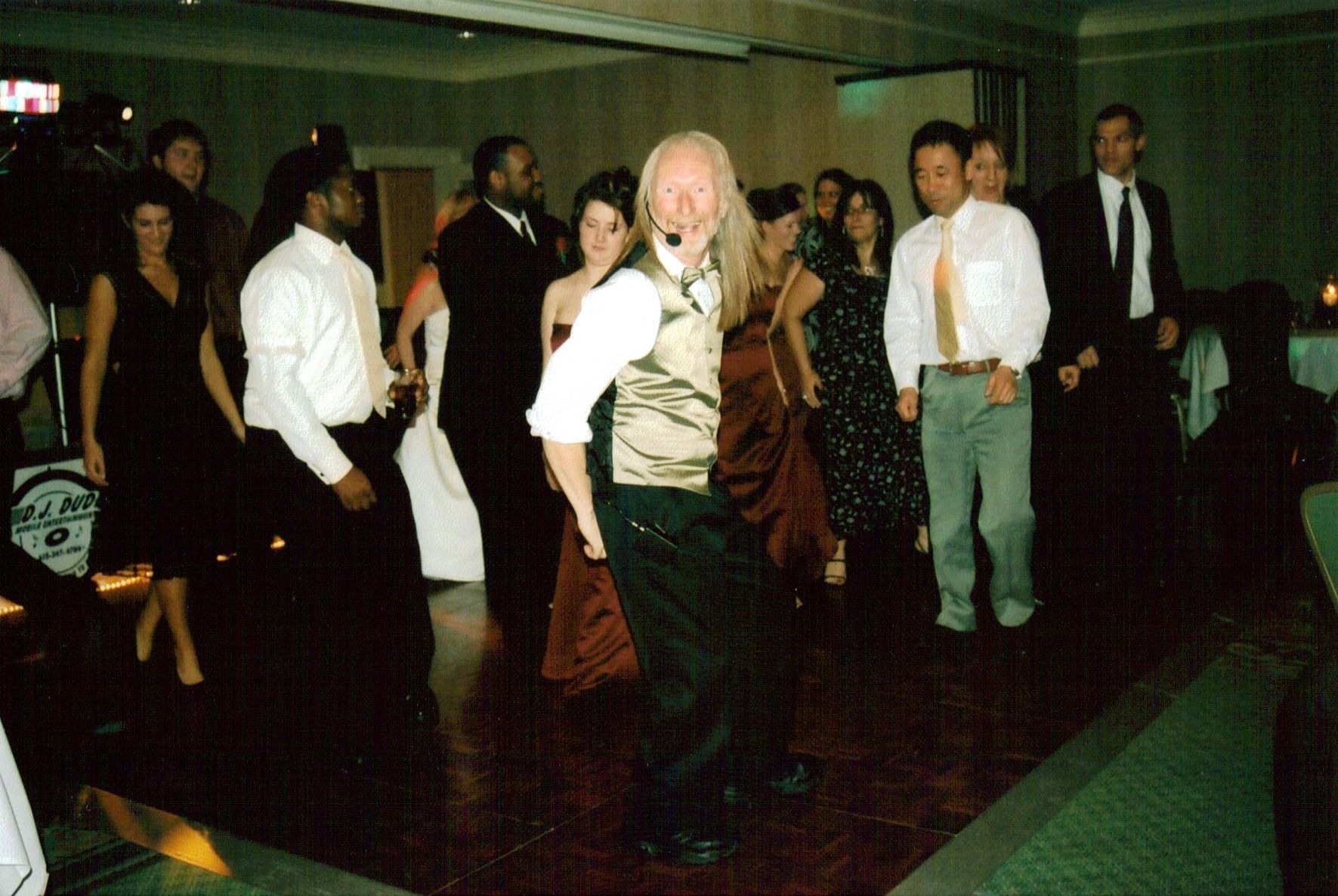 People dancing at a dimly lit indoor party, with one in a blond wig and brown vest in the center
