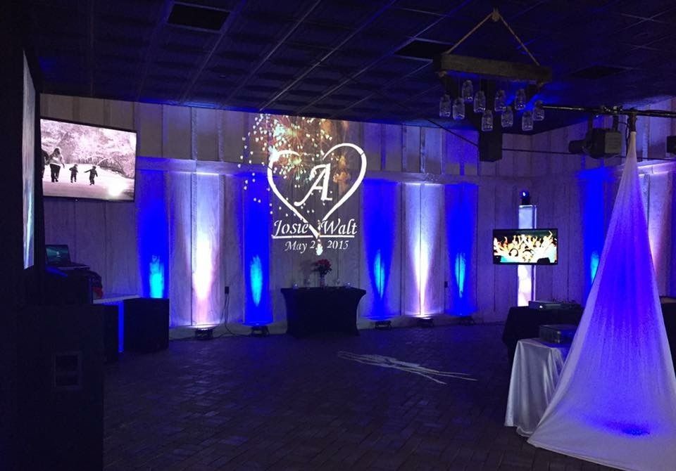 Blue-lit event hall with draped stage backdrop, central monogram logo, and small display screens on both sides