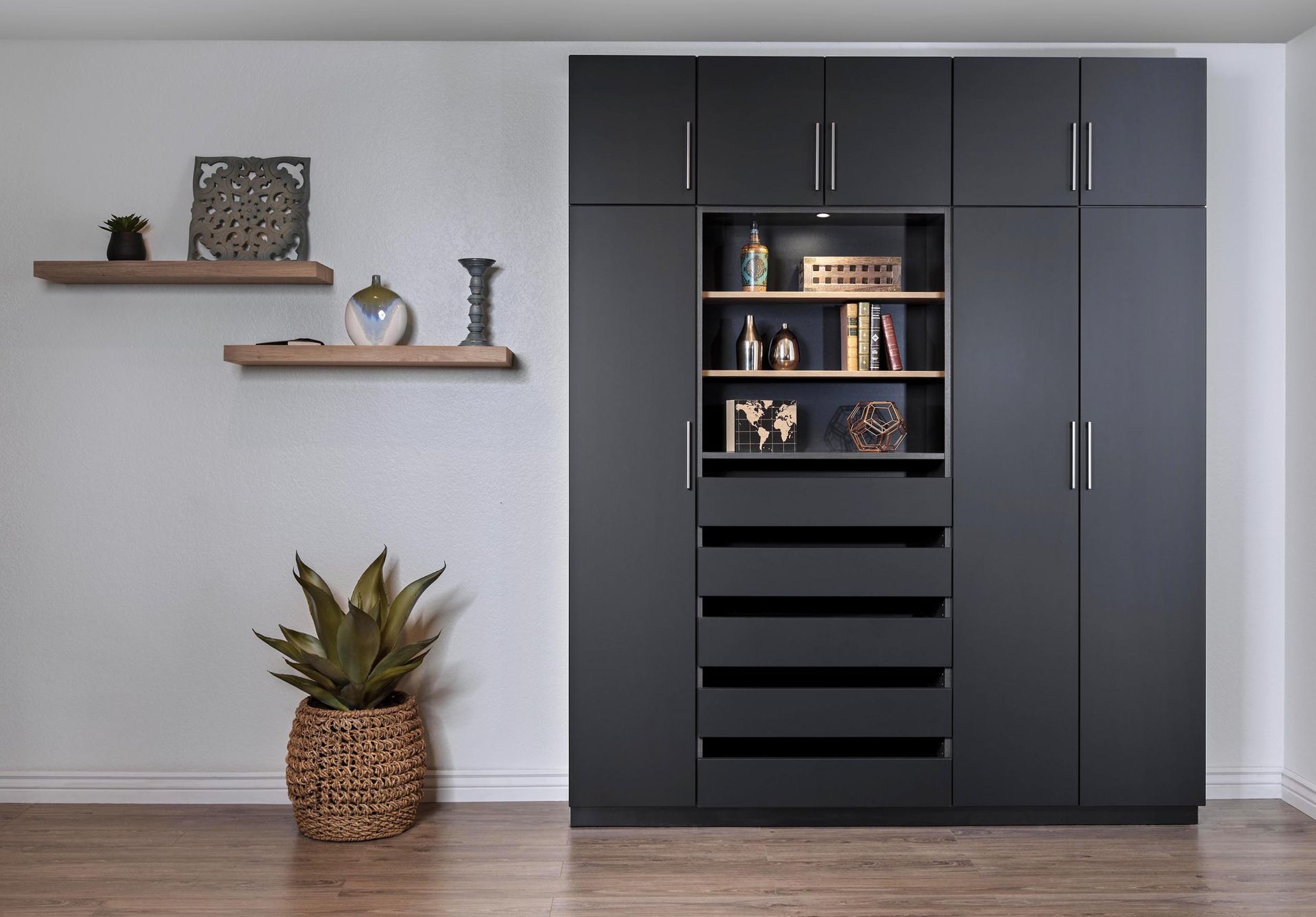 Dark gray wardrobe with built-in shelves and drawers, beside floating shelves and a potted plant in a light room.