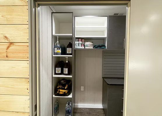 Pantry with tall open shelves holding cleaning supplies and food. Gray cabinets, white walls, and wood siding.