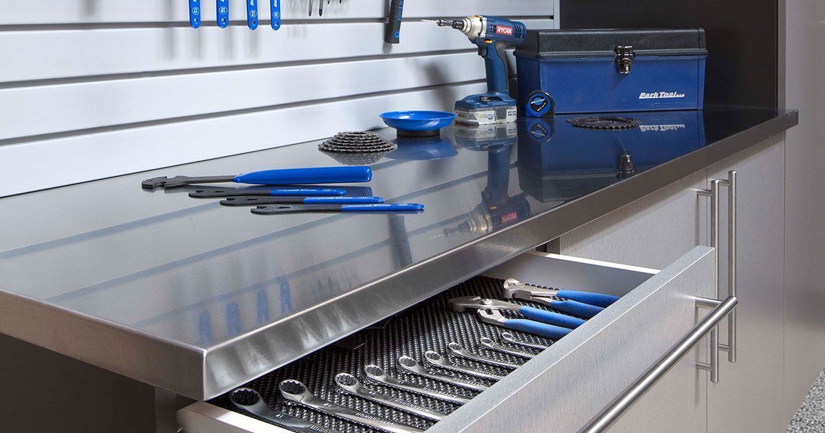 Stainless steel workbench with tools: open drawer, blue tools, toolbox, and slatted wall.