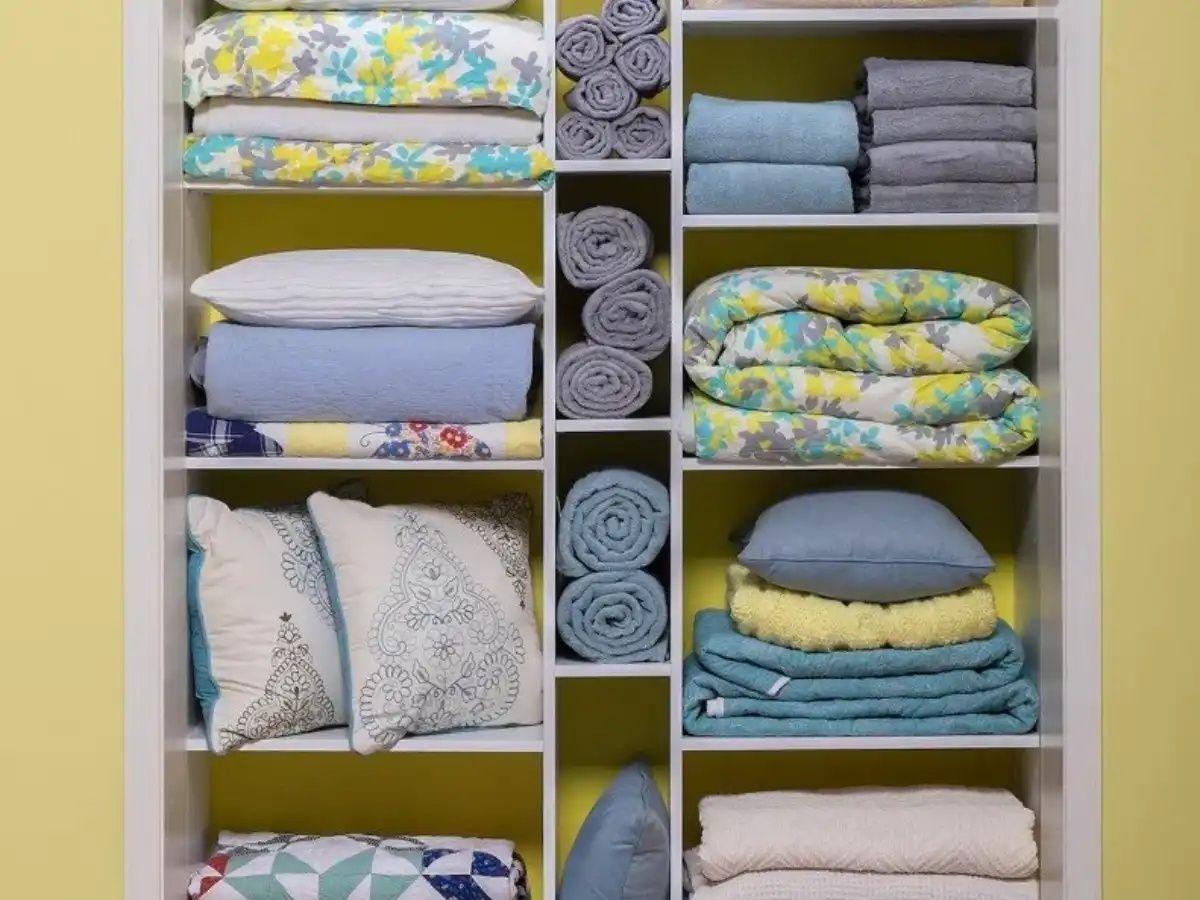White shelving unit filled with neatly stacked bedding, towels, and pillows; yellow backdrop.