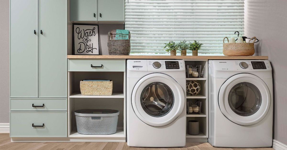 Laundry room with sage green cabinets, a washing machine, a dryer, and decorative baskets and plants.