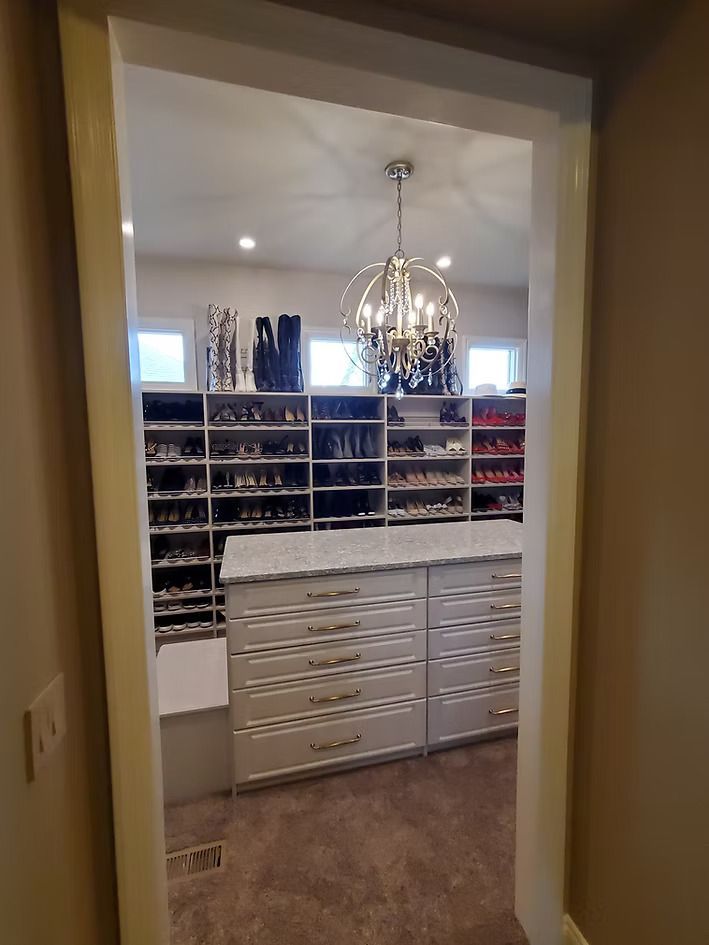A walk-in closet with a central island. Shoe shelves line the back wall, a chandelier hangs from the ceiling.