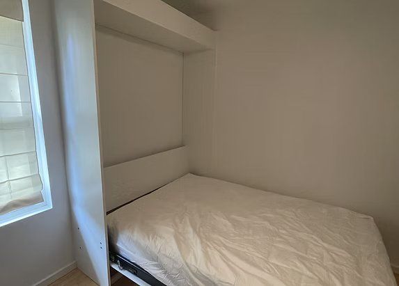 A small white bedroom with a bed, a window, and a built-in headboard.