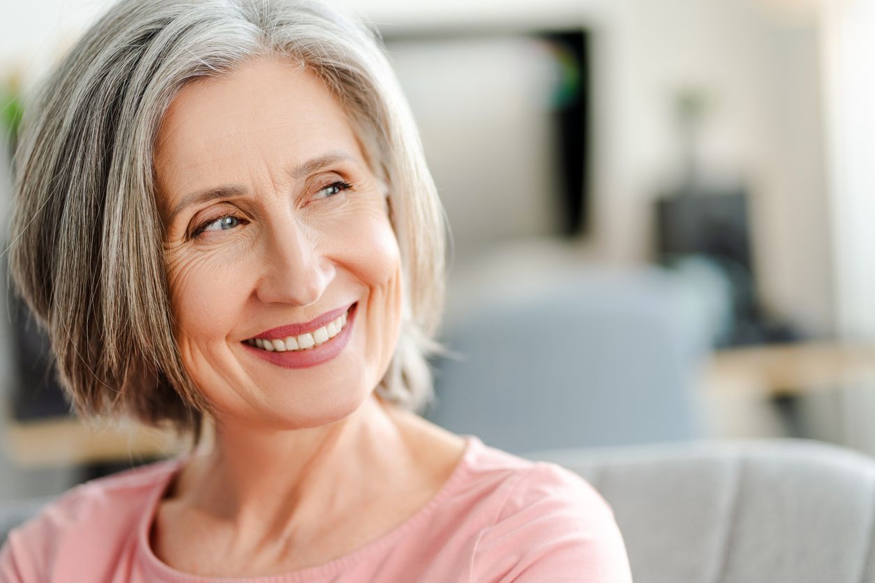 A woman with gray hair is smiling while sitting on a couch.