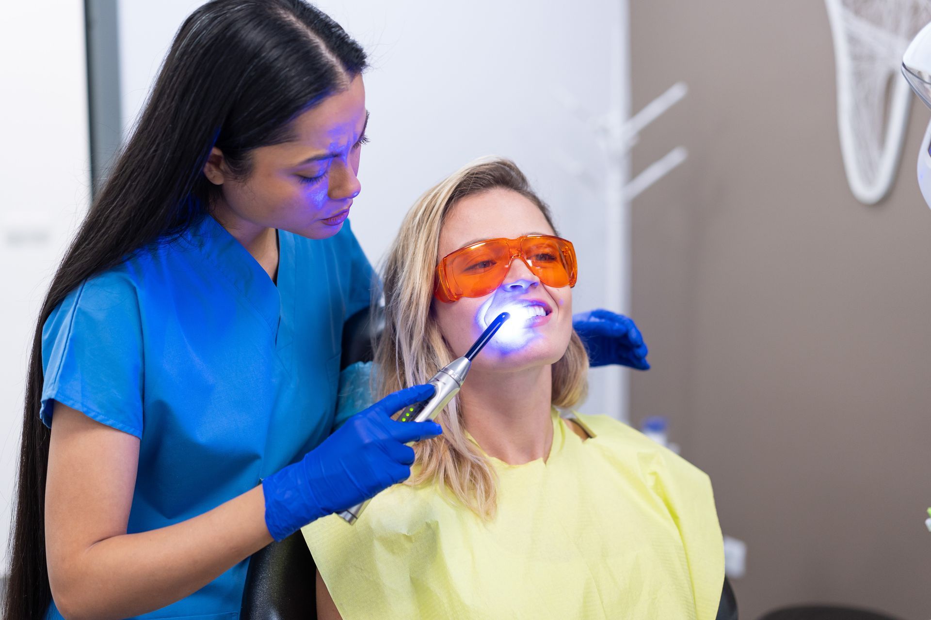 Dentist using a blue light on patient's teeth; patient wears protective glasses.