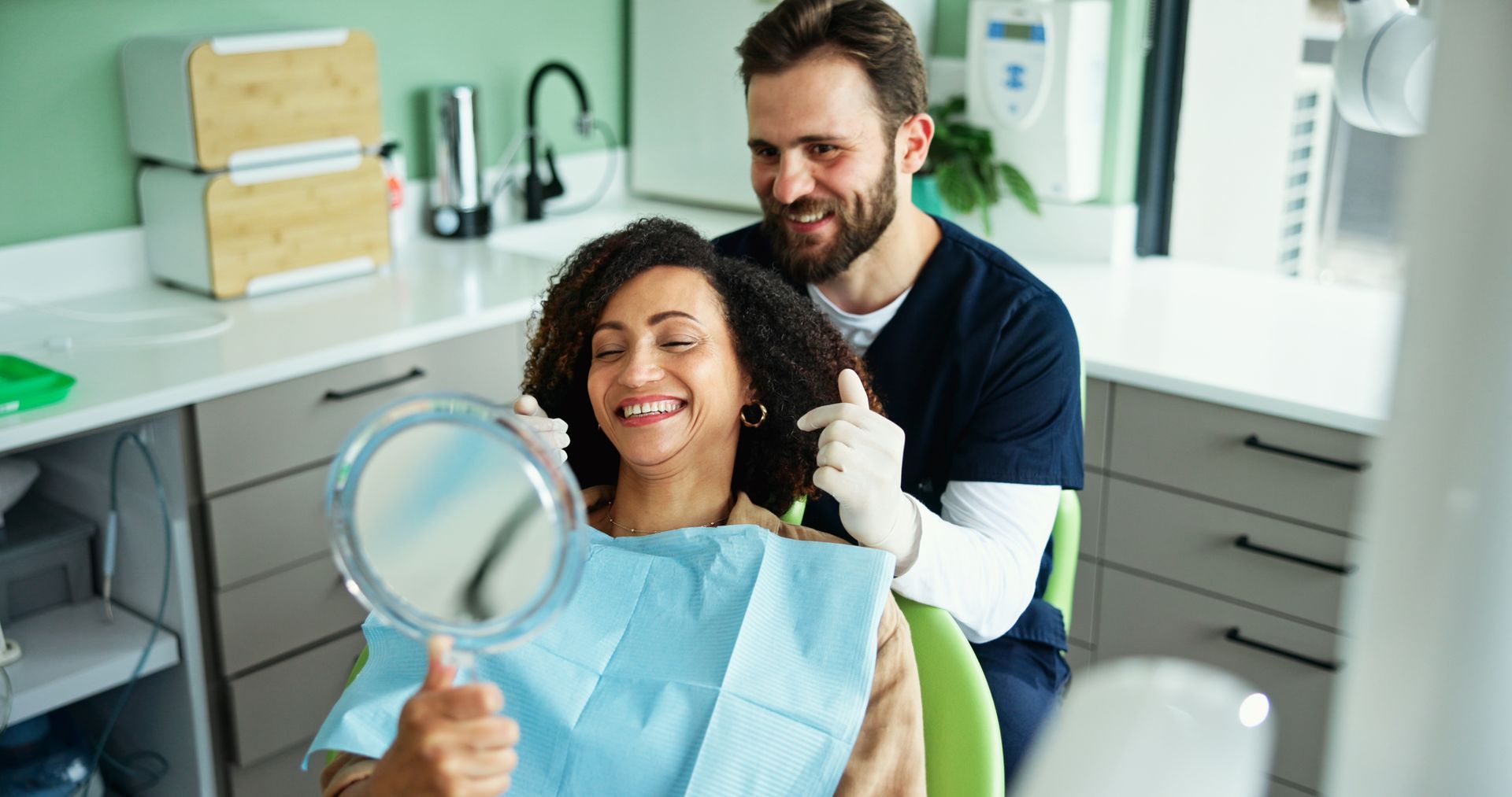 Dentist showing a smiling patient her teeth in a mirror. Green walls, light setting.