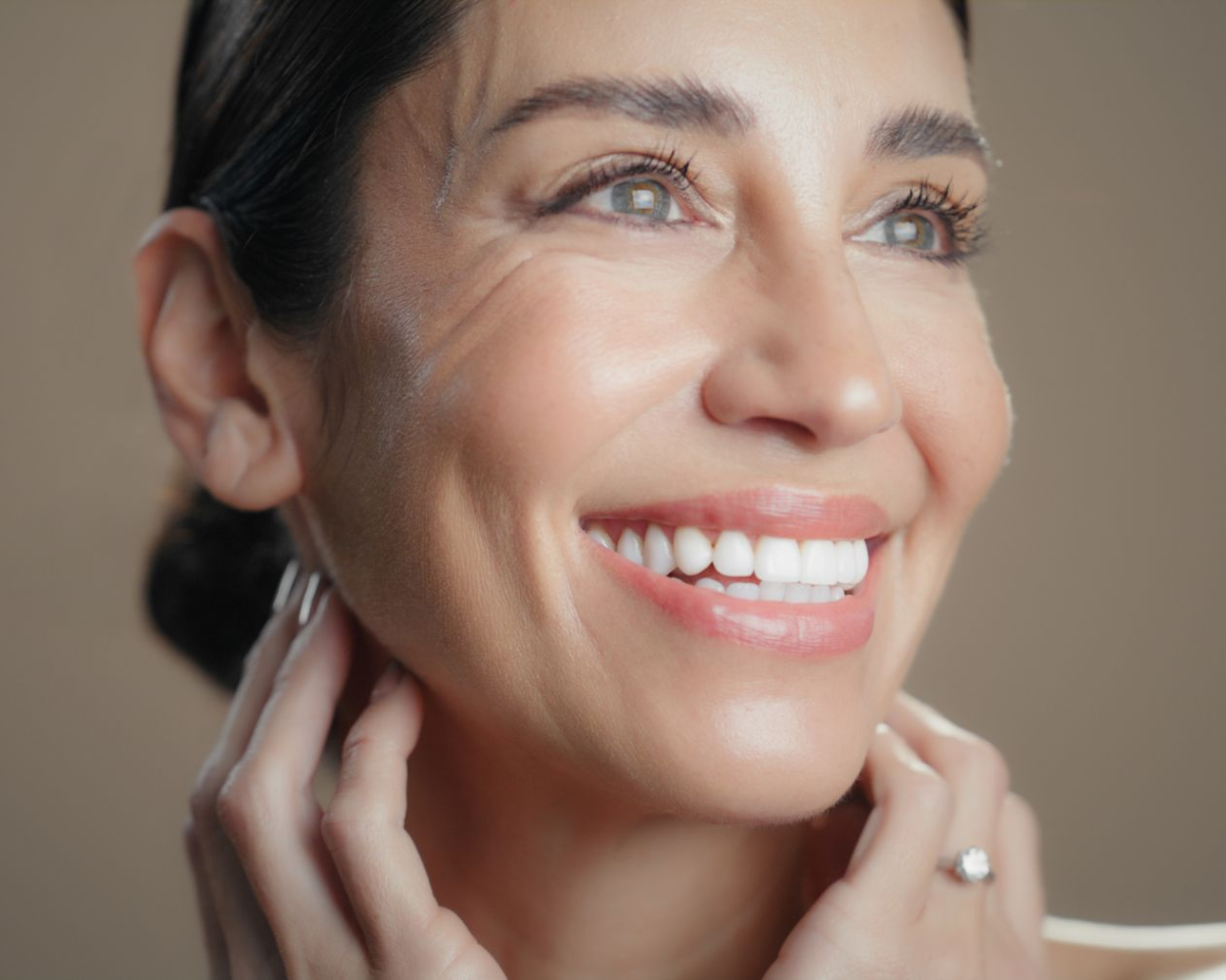 Woman smiling, looking upwards, hands at her neck; beige background.
