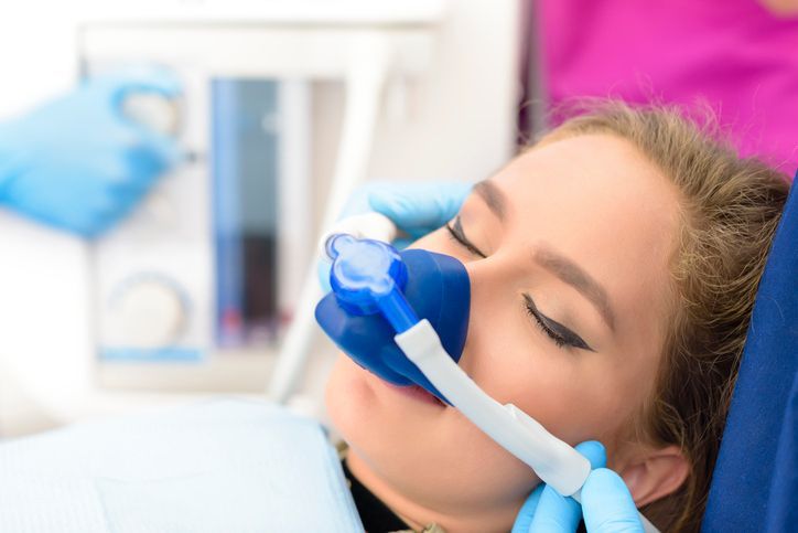 A person receiving nitrous oxide sedation through a nasal mask in a dental chair, with a gloved hand adjusting the unit.