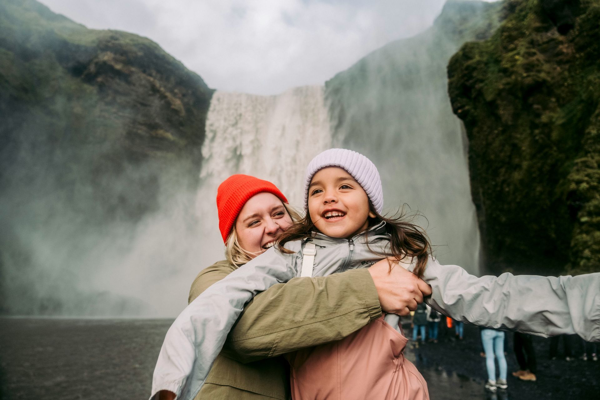 A woman is hugging a little girl in front of a waterfall.