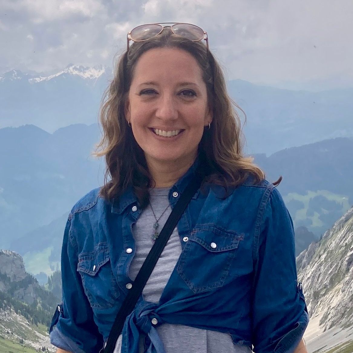 A woman in a blue shirt and sunglasses is standing on top of a mountain.